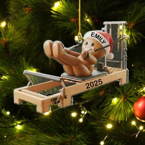 Op de afbeelding: Een kerstornament met een peperkoeken mannetje met een kerstmuts met de naam "Emily" erop, die oefent op een Pilates reformer. Het ornament heeft het jaartal "2025". De achtergrond is een kerstboom.