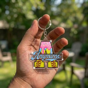 May include: A personalized softball keychain with a pink and blue design. The keychain features the text "Anyname" in a stylized font, with a softball graphic at the bottom. The keychain is held in a hand.