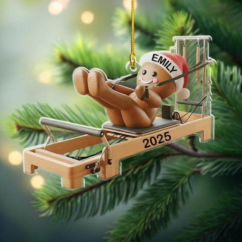 Op de afbeelding: Een kerstornament met een peperkoeken mannetje met een kerstmuts met de naam "Emily" en die oefent op een Pilates reformer. Het ornament is bruin en beige, met het jaartal "2025" op de reformer. Het hangt aan een gouden draad.