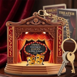 May include: A theater-themed keychain featuring a stage design with red curtains and gold masks. The keychain includes a gold chain, ring, and two keys. The text on the stage reads "EMMA Starring as JODAN in Blue School 2025."