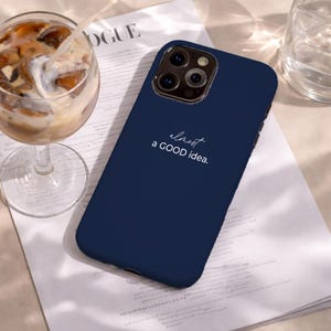 May include: A navy blue phone case with the text "almost a GOOD idea." The case is on a white surface next to a glass of iced coffee and a glass of water. The word "VOGUE" is visible in the background.