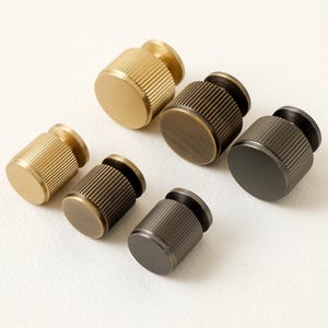 Könnte beinhalten: Sechs zylindrische Schrankknöpfe in verschiedenen Metallausführungen: Gold, Bronze und Dunkelgrau. Jeder Knopf hat einen geriffelten Körper und eine flache Ober- und Unterseite, geeignet für Möbel oder Dekoration.