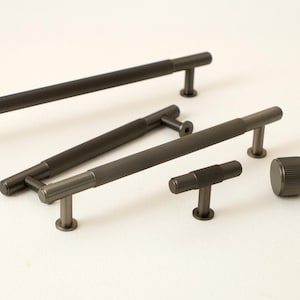 Pode incluir: Uma coleção de ferragens de armário cinza escuro, incluindo puxadores de barra e um botão. Os puxadores têm um design texturizado e canelado. As ferragens são feitas de metal e são mostradas em um fundo branco. Os puxadores de barra variam em comprimento.