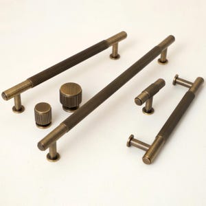 Op de afbeelding: Een verzameling antieke messing kastbeslagen, waaronder lange stanggrepen en ronde knoppen. De grepen hebben een getextureerd, geribbeld ontwerp en de knoppen zijn cilindrisch. De beslagen worden gepresenteerd tegen een witte achtergrond, die de warme, metalen afwerking benadrukt.