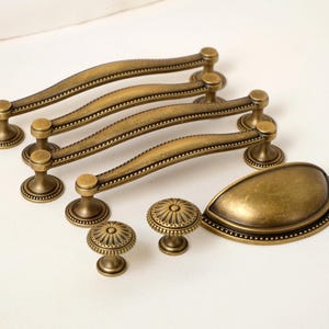 Puede incluir: Un conjunto de herrajes de gabinete de latón antiguo, que incluye cuatro manijas curvas, dos perillas redondas y un tirador en forma de lágrima. Los herrajes presentan un diseño detallado con acentos decorativos y un acabado cálido y envejecido.