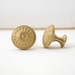 Puede incluir: Dos tiradores dorados para armarios con diseño floral. Los tiradores tienen una cara redonda y texturizada con un botón central y un borde con cuentas. Un tirador está en posición vertical, el otro de lado, mostrando el vástago. Cada tirador mide unos 4 cm de diámetro.