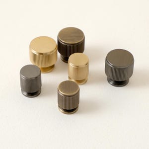 Peut inclure: Ensemble de six boutons de meubles m&eacute;talliques aux finitions vari&eacute;es, or, bronze et gris fonc&eacute;. Chaque bouton a une forme cylindrique avec une texture stri&eacute;e sur les c&ocirc;t&eacute;s et un dessus plat. Mesures en centim&egrave;tres.