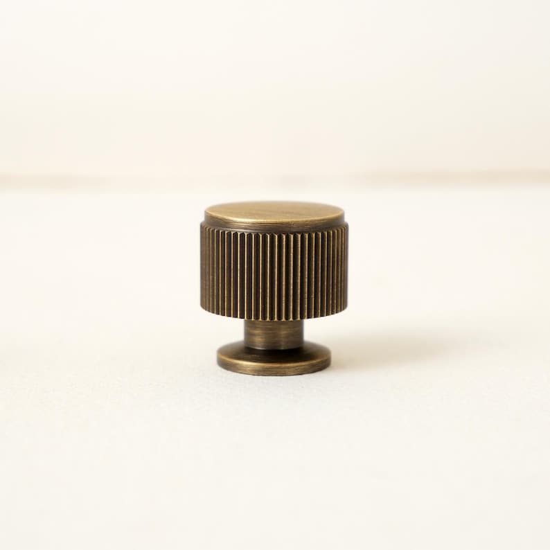Peut inclure: Un bouton de meuble en bronze avec un design cylindrique. Le dessus et la base sont plats, tandis que la section centrale pr&eacute;sente des d&eacute;tails verticaux et stri&eacute;s. Le bouton est con&ccedil;u pour une prise en main facile et ajoute une touche de style vintage aux meubles.