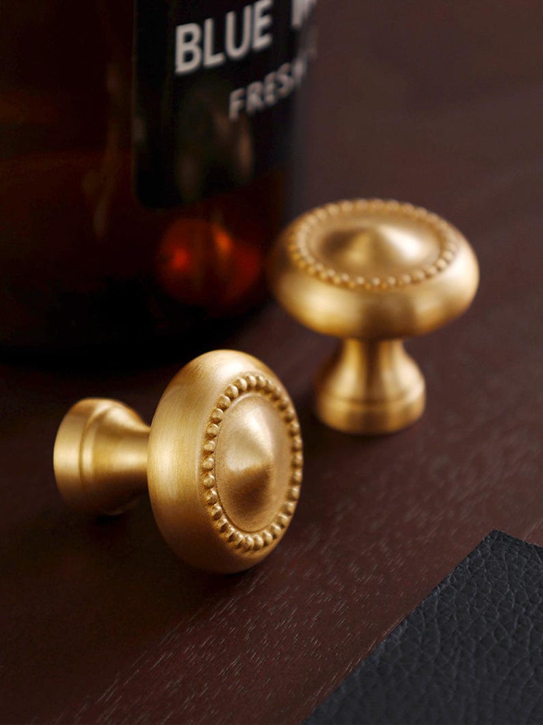 May include: Two brass cabinet knobs with a round, beaded edge design. The knobs have a mushroom-shaped base and a conical center. The knobs are displayed on a dark brown surface. The image also shows a blurred brown bottle with the text "BLUE FRESH".