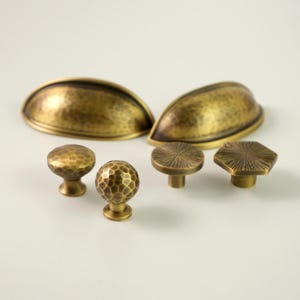 May include: A selection of antique brass cabinet hardware, featuring two cup pulls, two round knobs, and two geometric knobs. The hardware has a hammered texture and a warm, aged finish, suitable for kitchen or furniture renovations.