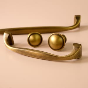 May include: A selection of antique brass cabinet hardware, comprising two curved handles and two round knobs. The hardware features a warm, aged finish, indicative of a vintage or classic style. The pieces are displayed against a neutral backdrop, accentuating their design.