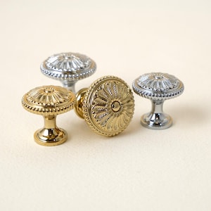May include: Four decorative cabinet knobs in gold and silver tones. The knobs feature a floral design with intricate detailing. The knobs are made of metal and have a polished finish. The knobs are of varying sizes and shapes.