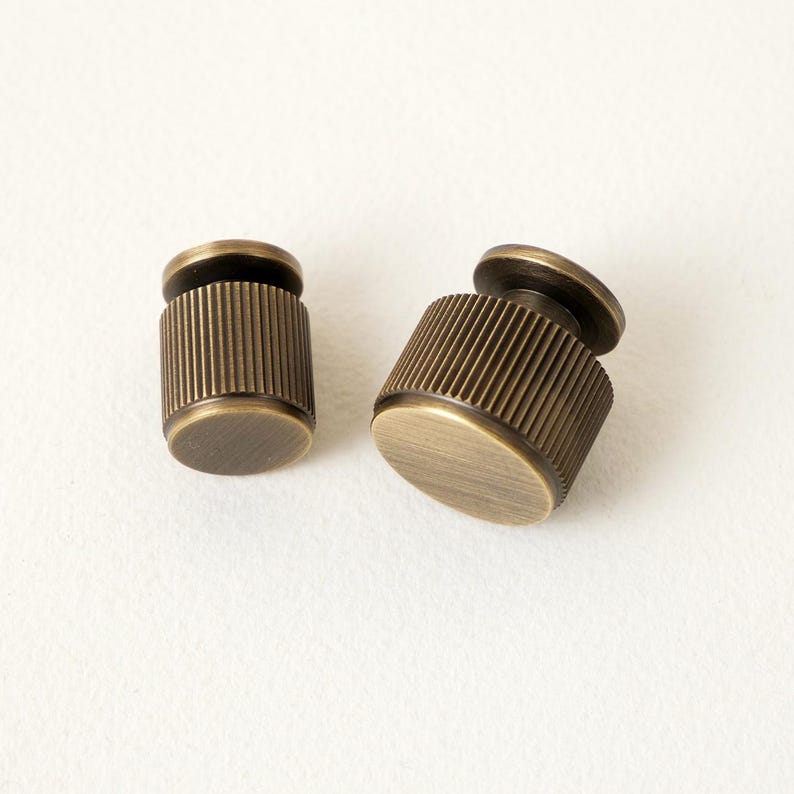 Peut inclure: Deux boutons de meuble en laiton antique. Chaque bouton pr&eacute;sente un corps cylindrique stri&eacute; et une base circulaire plate, avec un petit dessus arrondi. Les boutons sont pr&eacute;sent&eacute;s sur un fond blanc, mettant en valeur leur finition m&eacute;tallique vieillie.