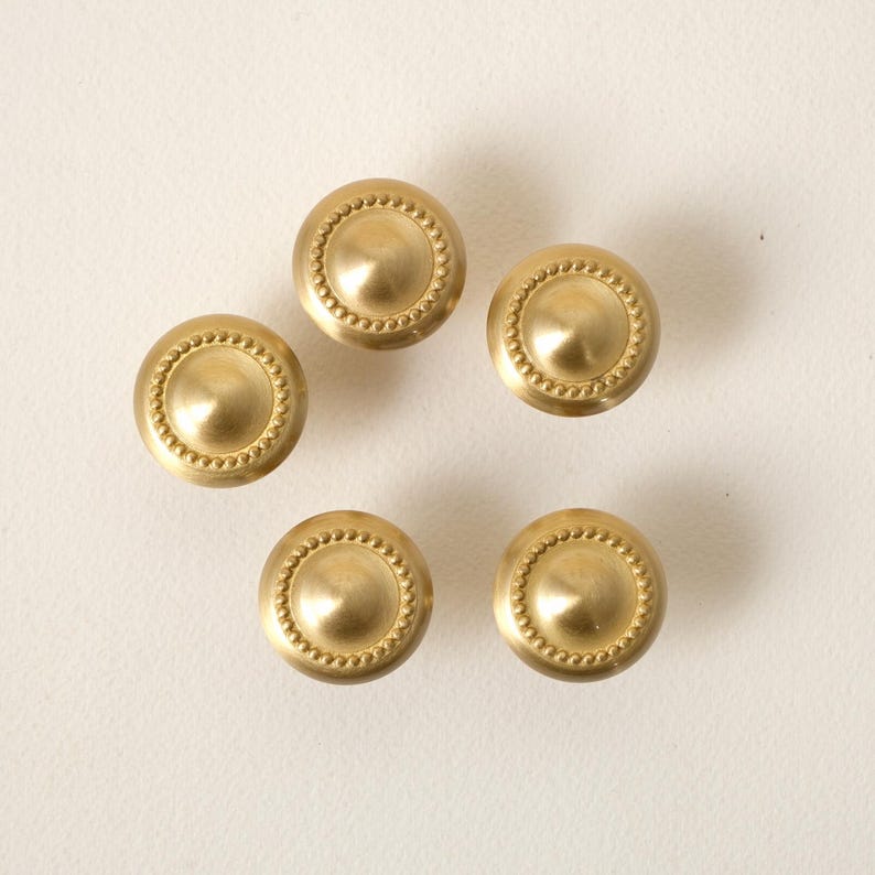May include: Five brass cabinet knobs with a circular design. Each knob has a central, slightly recessed area surrounded by a beaded border. The knobs are a warm, golden color and are arranged on a white surface.