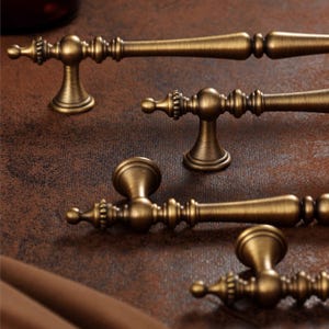 May include: Close-up of several antique brass cabinet pulls. The pulls have a detailed design with a central knob and decorative ends. The hardware is a warm, aged brass colour, and the background is a textured brown.