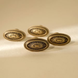 Könnte beinhalten: Vier ovale Schrankknöpfe aus antikem Messing. Jeder Knopf hat ein detailliertes Blumendesign in der Mitte, umgeben von einem Perlenrand. Die Knöpfe haben eine warme, gealterte Oberfläche, die einen Vintage-Stil suggeriert. Ideal für Möbel.