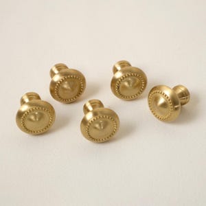 May include: Five brass cabinet knobs with a rounded, domed design. Each knob features a central, slightly raised disc surrounded by a ring of small, decorative beads. The knobs have a warm, golden hue and are arranged on a neutral background.