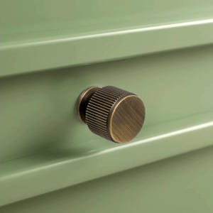 Peut inclure: Gros plan sur un tiroir vert sauge avec un bouton rond en laiton antique. Le bouton a une texture stri&eacute;e et une finition m&eacute;tallique chaude. Le devant du tiroir est lisse et peint en vert.