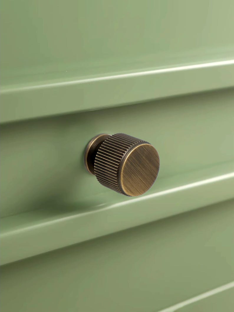 Peut inclure: Gros plan sur un tiroir vert sauge avec un bouton rond en laiton antique. Le bouton a une texture stri&eacute;e et une finition m&eacute;tallique chaude. Le devant du tiroir est lisse et peint en vert.