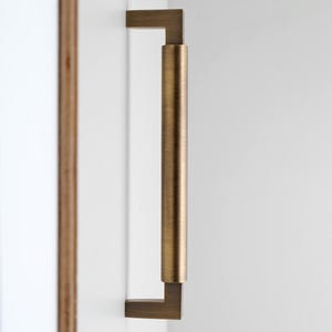 Peut inclure: Une poignée de meuble de couleur bronze fixée sur une surface blanche. La poignée a une base rectangulaire et une section centrale cylindrique. Ce matériel moderne convient aux armoires de cuisine ou de salle de bain. Dimensions en centimètres.