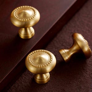 May include: Three brass cabinet knobs with a round, beaded design on a dark brown surface. The knobs have a classic, elegant look, suitable for furniture or cabinetry. The knobs are made of brass.
