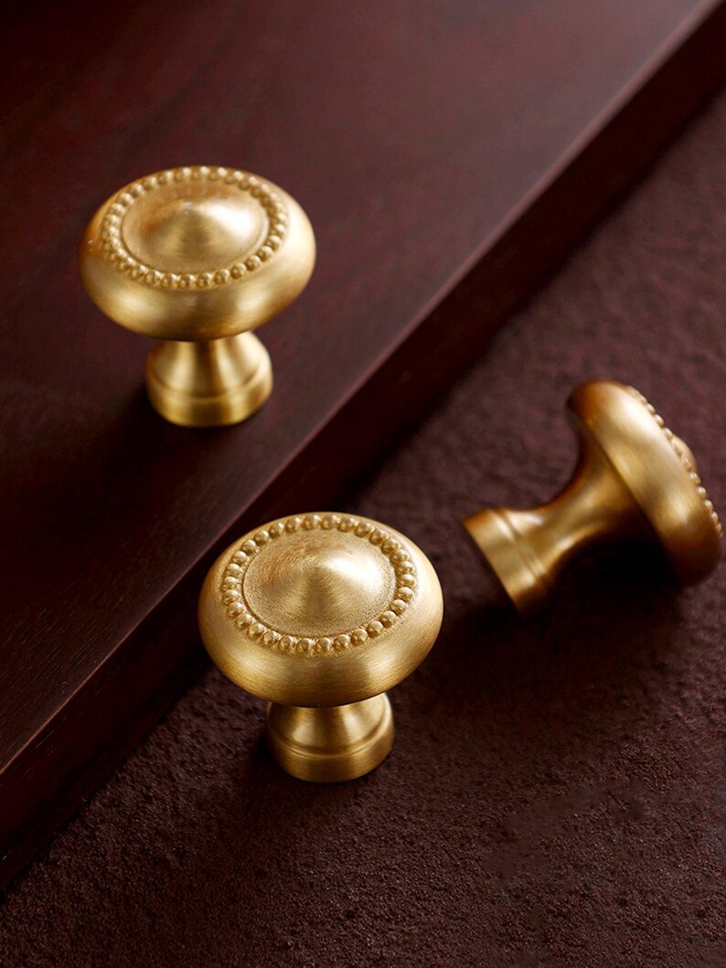 May include: Three brass cabinet knobs with a round, beaded design on a dark brown surface. The knobs have a classic, elegant look, suitable for furniture or cabinetry. The knobs are made of brass.