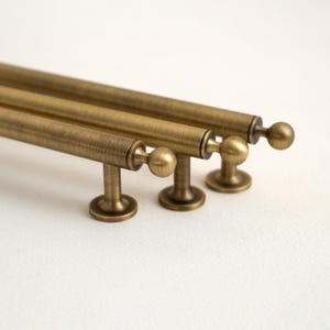 Peut inclure: Gros plan sur trois poignées de meubles en laiton antique. Chaque poignée présente une barre cylindrique avec une extrémité en forme de boule et une base circulaire. La quincaillerie a une finition chaude et vieillie, suggérant une esthétique vintage ou rustique.
