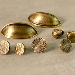 May include: A collection of brass cabinet hardware, including cup pulls and various knob styles. The hardware has a hammered texture and a warm, antique gold finish. The knobs include round, hexagonal, and spherical designs.
