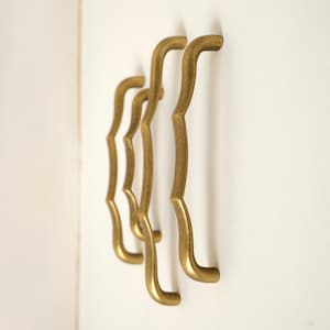 Puede incluir: Cuatro tiradores dorados para armarios con un diseño en forma de corchete están montados sobre una superficie blanca. Los tiradores tienen una forma curva con un patrón ondulado decorativo. El herraje es probablemente para un armario o un cajón.