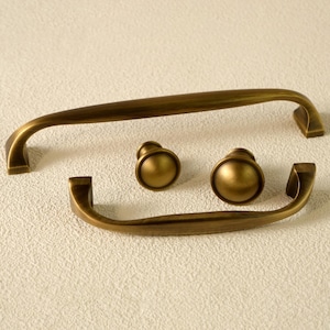 May include: Set of antique brass cabinet hardware, including two curved handles and two round knobs. The hardware has a warm, aged finish, and is designed for furniture restoration or new builds.