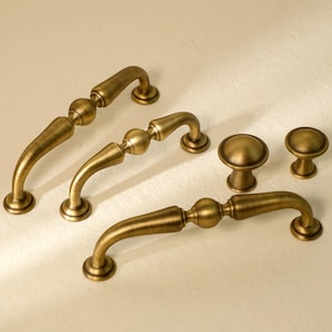 May include: A collection of antique brass cabinet hardware, including three curved pull handles and two round knobs. The handles feature a decorative center and curved ends, while the knobs have a simple, rounded design. The hardware has a warm, aged finish.