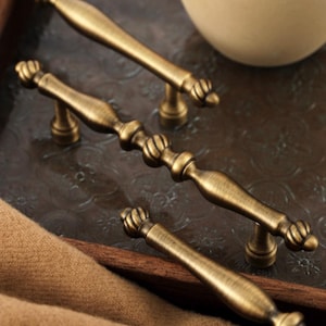 Op de afbeelding: Vier antieke messing kastgrepen met een decoratief ontwerp. De grepen zijn gerangschikt op een donkerbruin dienblad met een bruin textiel op de voorgrond. De grepen hebben een klassieke, vintage uitstraling.