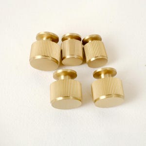 Peut inclure: Six boutons de meuble en laiton avec un corps cylindrique et un petit dessus circulaire. Les boutons ont une texture stri&eacute;e sur le corps et une finition lisse et polie. Les boutons sont dispos&eacute;s sur une surface blanche.