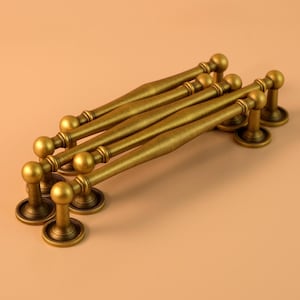 Peut inclure: Six poignées de meubles en laiton antique avec une finition vieillie. Chaque poignée présente un bouton arrondi à chaque extrémité et un centre légèrement incurvé. Les poignées sont disposées en diagonale sur un fond neutre.