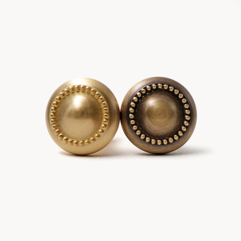 May include: Two round cabinet knobs. One is a bright gold color, and the other is a darker bronze color. Both knobs have a central, raised, circular design surrounded by a ring of small, evenly spaced, raised dots. The knobs are likely made of metal.