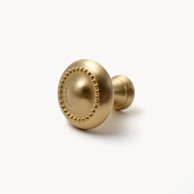 May include: A gold-colored cabinet knob with a round, beaded design around the edge. The knob has a smooth, matte finish and a small, cylindrical base. The hardware is likely for furniture or cabinetry.