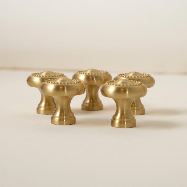 May include: Five brass cabinet knobs with a mushroom-shaped design. Each knob features a rounded top with a beaded edge and a cylindrical base. The knobs are a warm gold color and are arranged on a neutral background.