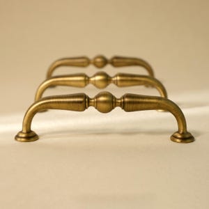 May include: Three antique brass cabinet pulls arranged in a row. Each pull has a curved handle with a central decorative sphere and rounded ends. The pulls are made of a warm, metallic brass.