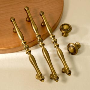 Könnte beinhalten: Satz antiker Messing-Schrankbeschläge, bestehend aus vier Griffen und zwei Knöpfen. Die Griffe haben ein dekoratives, verziertes Design mit geschwungener Form. Die Knöpfe sind rund mit einem perlenbesetzten Rand. Die Beschläge liegen auf einer hellen Oberfläche.