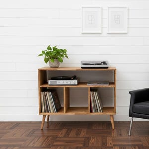 May include: A wooden entertainment unit with a record player, vinyl records, and a black leather armchair. The cabinet has a natural finish, and the chair has chrome legs. Two framed abstract art pieces hang on the white wall. The unit is 100cm wide.