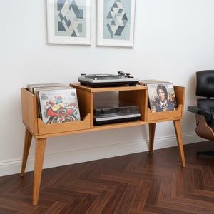 May include: A wooden record player console in a mid-century modern style. It features storage for vinyl records, a turntable, and a shelf for a second player. The room includes a black leather chair and two framed geometric prints. The console is approximately 120 cm long.
