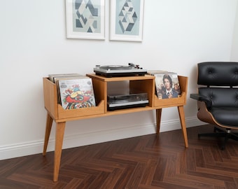 May include: A mid-century modern record player console made of light-colored wood. The console has two storage compartments for vinyl records, a turntable on top, and a shelf for a second record player. The room has a black leather chair and two framed geometric art prints.