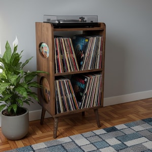 Op de afbeelding: Een platenspelerstandaard in mid-century modern stijl van donker hout. De standaard heeft twee planken gevuld met vinylplaten en een platenspeler bovenop. Links van de standaard staat een potplant.