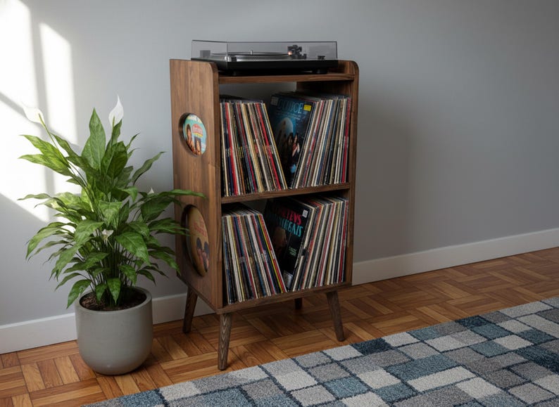 Op de afbeelding: Een platenspelerstandaard in mid-century modern stijl van donker hout. De standaard heeft twee planken gevuld met vinylplaten en een platenspeler bovenop. Links van de standaard staat een potplant.