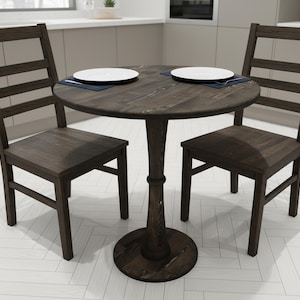 Puede incluir: Una mesa de comedor redonda de color marrón oscuro con dos sillas a juego. La mesa tiene una base de pedestal y está decorada con dos platos blancos sobre manteles individuales azules. Las sillas tienen respaldos de listones y asientos cuadrados. Los muebles son de madera.
