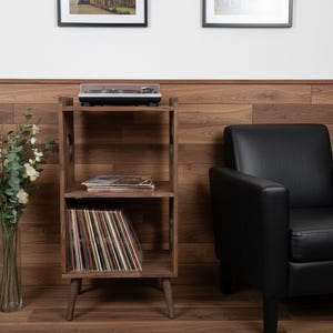 Op de afbeelding: Een houten platenspelerstandaard met drie niveaus. Bovenop staat een platenspeler, in het midden een stapel platen en onderaan vinylplaten. Rechts staat een zwarte leren fauteuil.