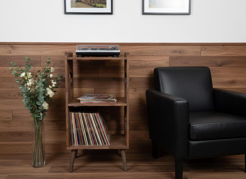 Op de afbeelding: Een houten platenspelerstandaard met drie niveaus. Bovenop staat een platenspeler, in het midden een stapel platen en onderaan vinylplaten. Rechts staat een zwarte leren fauteuil.