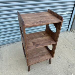 Op de afbeelding: Een houten plank met drie niveaus en een donkerbruine afwerking. De planken zijn rechthoekig en de zijkanten hebben grote, cirkelvormige uitsparingen. De plank staat op vier korte poten. De bovenste plank heeft een verhoogde achterkant.