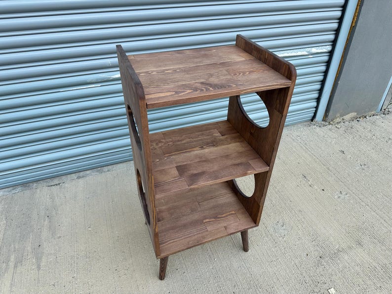 Op de afbeelding: Een houten plank met drie niveaus en een donkerbruine afwerking. De planken zijn rechthoekig en de zijkanten hebben grote, cirkelvormige uitsparingen. De plank staat op vier korte poten. De bovenste plank heeft een verhoogde achterkant.