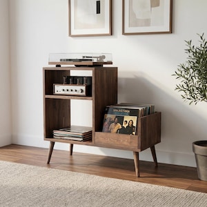 Könnte beinhalten: Ein Plattenspieler-Ständer im Mid-Century-Modern-Stil mit warmbraunem Finish. Der Ständer verfügt über einen Plattenspieler, einen Verstärker und Stauraum für Schallplatten. Daneben steht ein kleiner Olivenbaum in einem dunklen Topf.